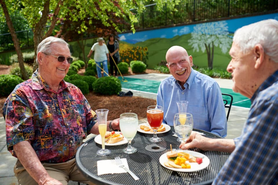 3 older gentlemen talking over snacks and drinks with couple playing shuffleboard in background